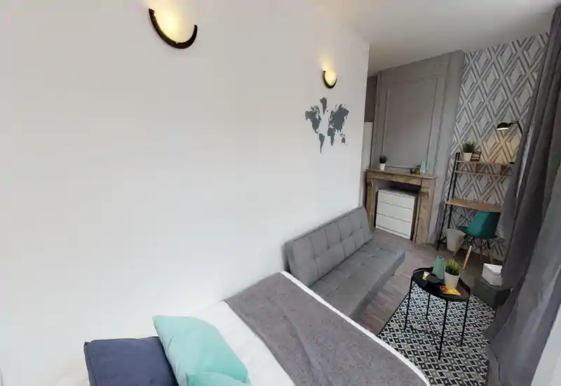 Chambre lumineuse et bien mise en scène avec lit visible, canapé convertible gris, tapis à motifs et petit coin bureau — ambiance moderne et confortable, idéale pour une personne ou un couple.