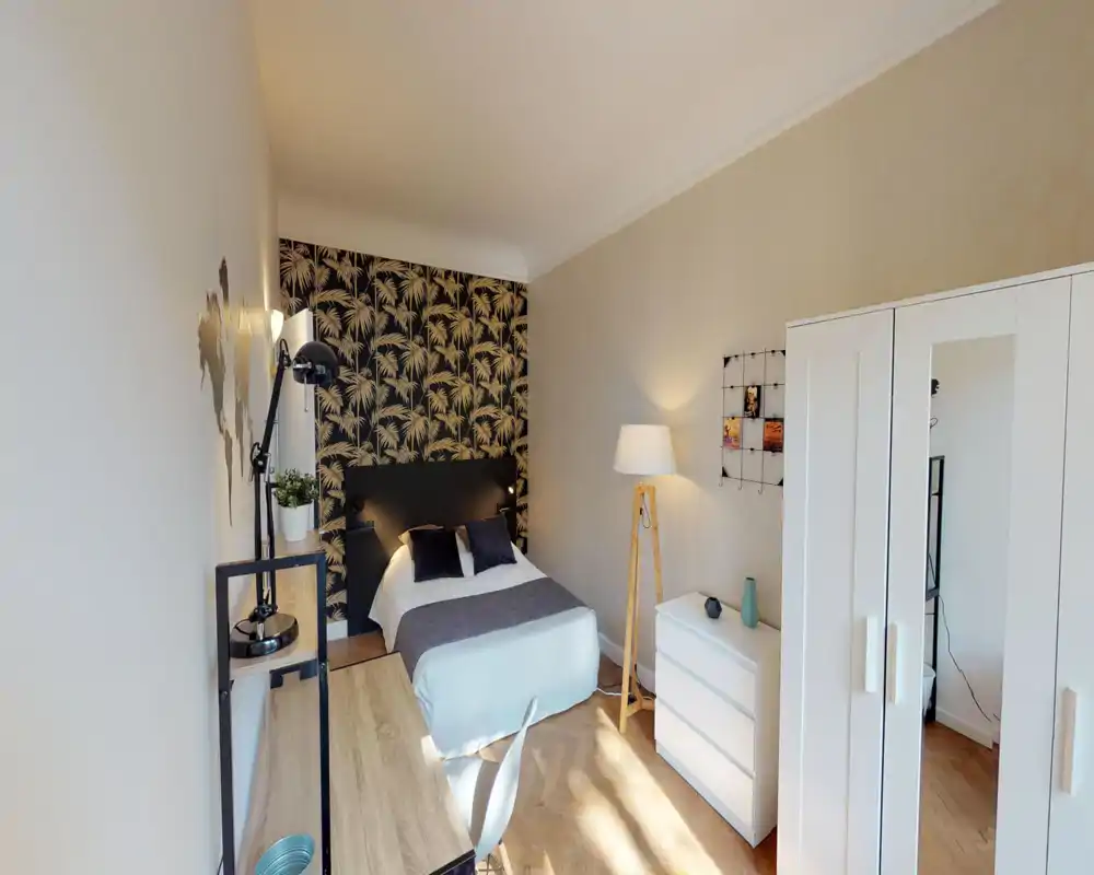 Bright, stylish bedroom with a centrally placed double bed, decorative palm-patterned accent wall, desk area and white wardrobe—well-lit and cozy for short or long stays.