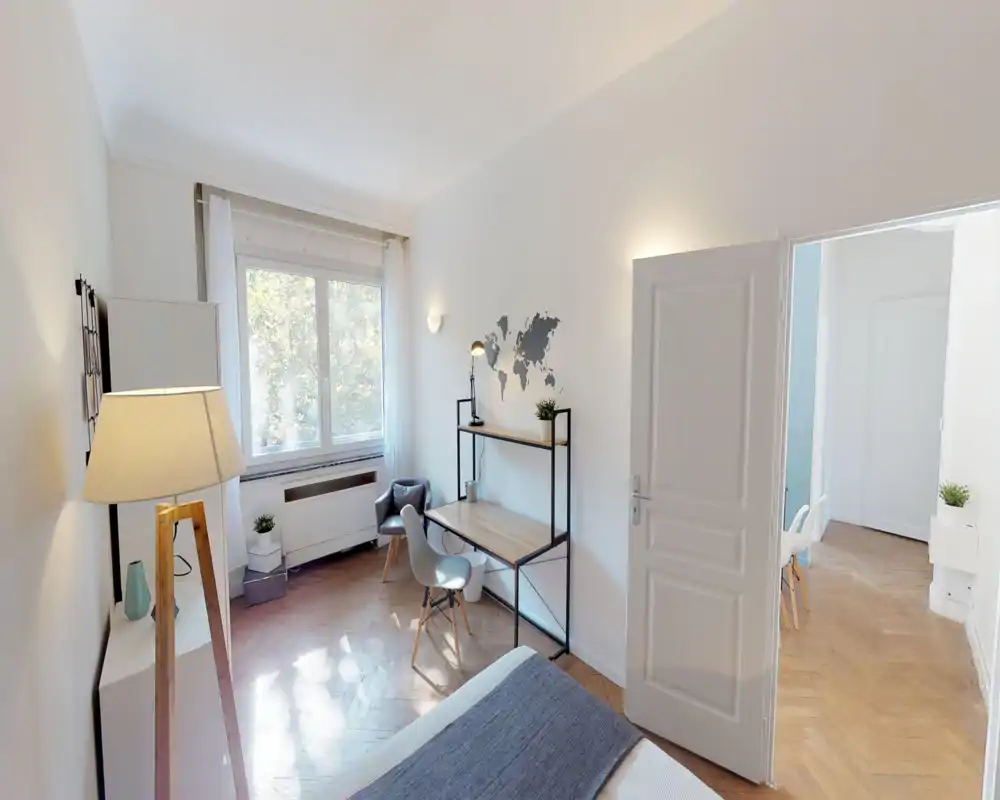 Bright, well-styled bedroom with a comfortable bed in the foreground, a work desk by the window and warm wooden floors that create a cozy, functional space.