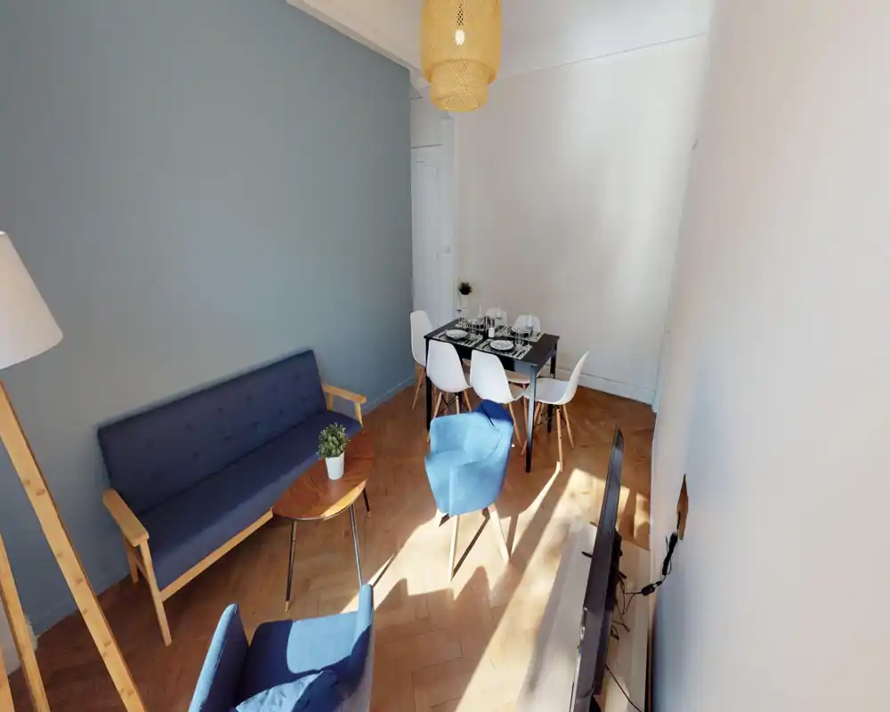 Bright, stylish living room with blue sofa and matching armchairs, a small coffee table and a dining area set for four. Natural light and warm wooden floors create a welcoming atmosphere.