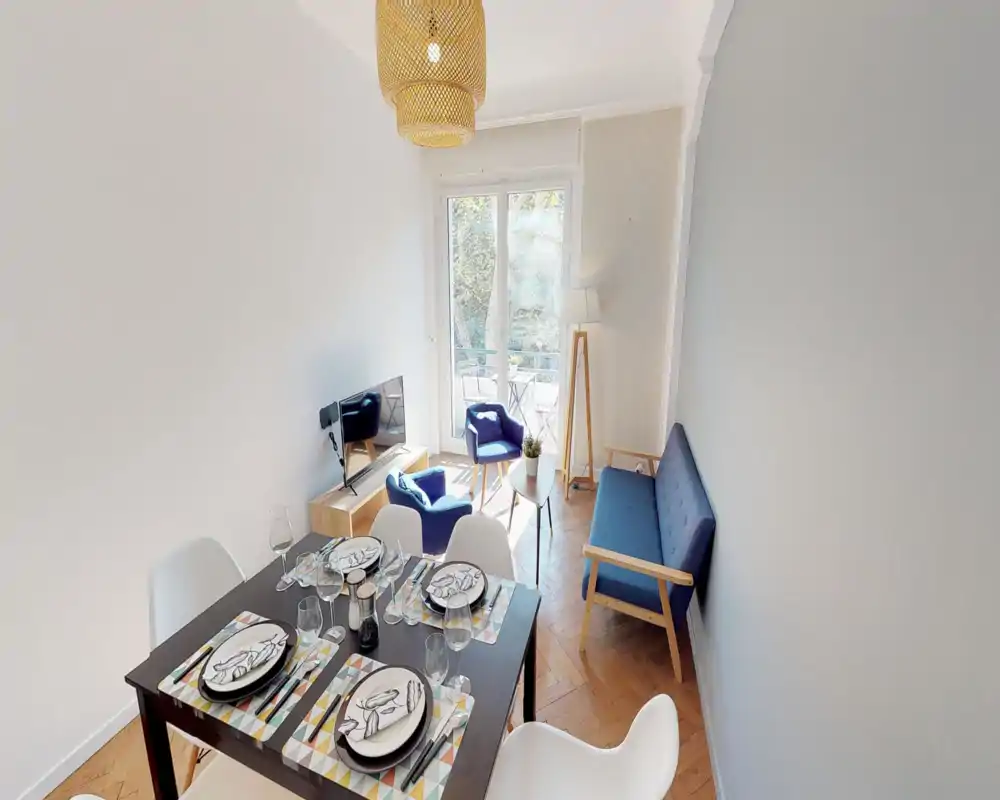 Bright, well-styled living room with a small dining table, blue sofa and armchairs, and a balcony door letting in natural light — ideal for a cozy apartment stay.
