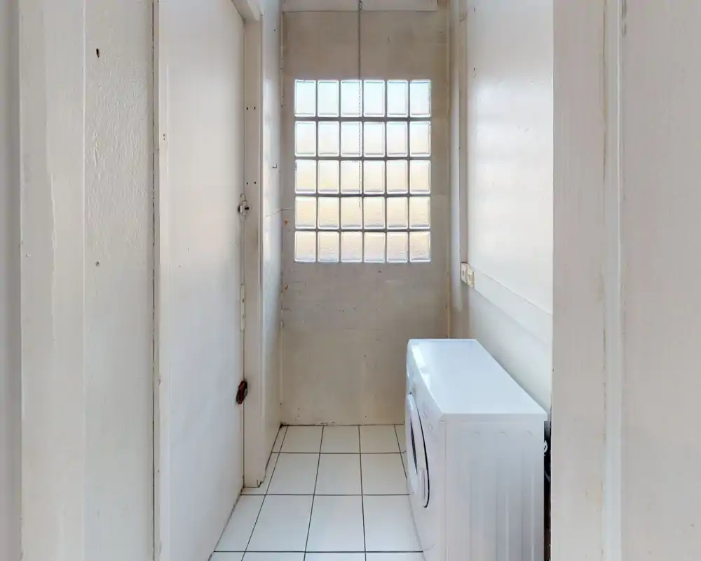 Small bright utility/laundry alcove with a front-loading washing machine, tiled floor and glass-block window providing natural light.