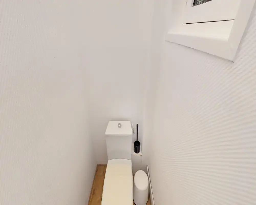 Compact toilet room with a standard cistern, toilet brush and a small waste bin; bright white textured walls and wood-effect floor create a clean, minimalist feel.