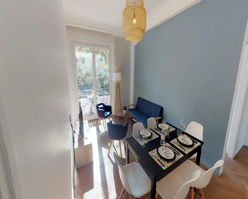Sunny, well-staged living and dining area with a sofa, armchairs and a dining table set for four; large glass doors open to a small balcony letting in abundant natural light.