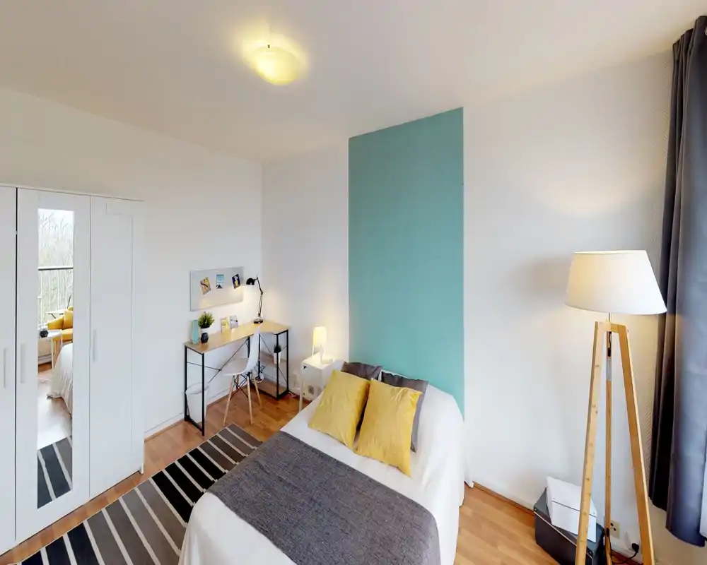 Bright, styled bedroom with a double bed against a teal accent panel, cozy yellow cushions, bedside lamps and a neat desk — ideal for both rest and work.