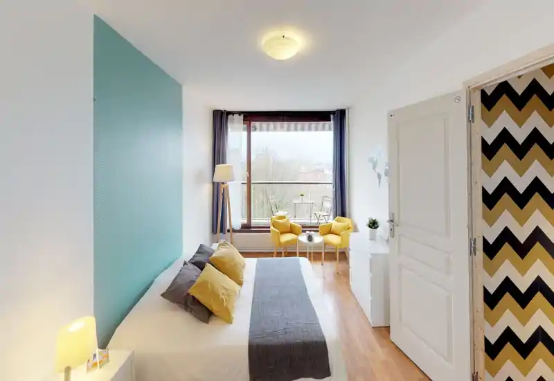 Bright, modern bedroom with a large window and balcony view; comfortable double bed with decorative pillows and a cozy seating area by the window.