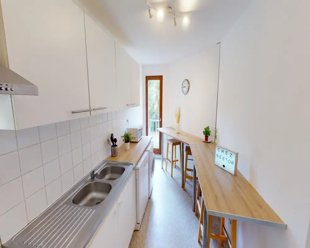 Narrow, modern galley kitchen with white cabinets, stainless sink, wooden countertops and a small breakfast bar by a balcony door. Bright and tidy space ideal for quick meals and light cooking.