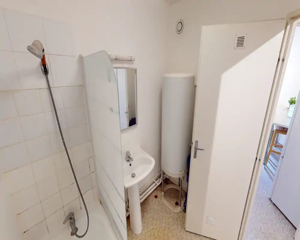 Compact bathroom with a shower stall, handheld showerhead, pedestal sink and a vertical water heater; light tiles and neutral walls create a simple, functional space.