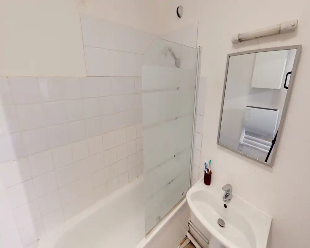 Compact bathroom with bathtub and wall-mounted partial glass screen, small sink with mirror and toothbrush holder; clean white tiles and good natural lighting.