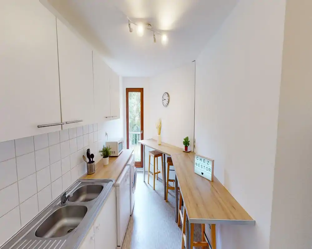 Bright, narrow kitchen with white cabinets, dual stainless sink and wooden counters forming a breakfast bar with stools — compact and well organized for short-term stays.