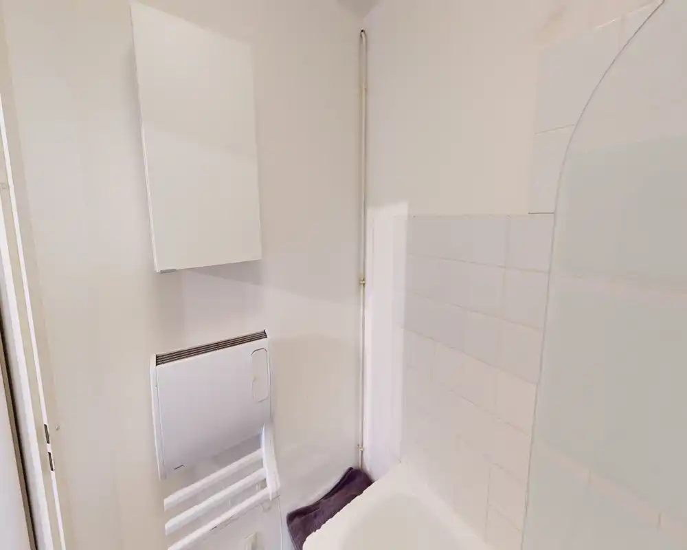 Compact bathroom corner with tiled walls, a bathtub edge, wall-mounted heater/radiator and a small storage cabinet. Bright, clean and practical layout.