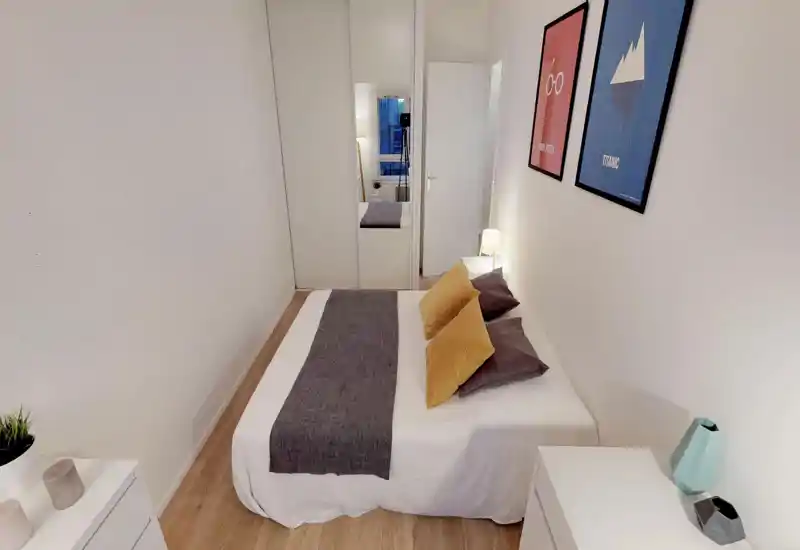 Camera da letto luminosa e ben arredata con letto matrimoniale, cuscini decorativi e copriletto, lampada da comodino e quadri a parete. Armadio con ante a specchio e pavimento in legno chiaro ampliano lo spazio.