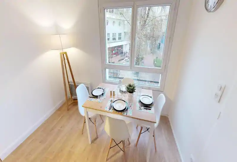 Zona pranzo luminosa e curata con tavolo in legno apparecchiato per quattro persone vicino a una grande finestra. Sedie dallo stile scandinavo e una lampada da terra rendono l'ambiente accogliente e arioso.