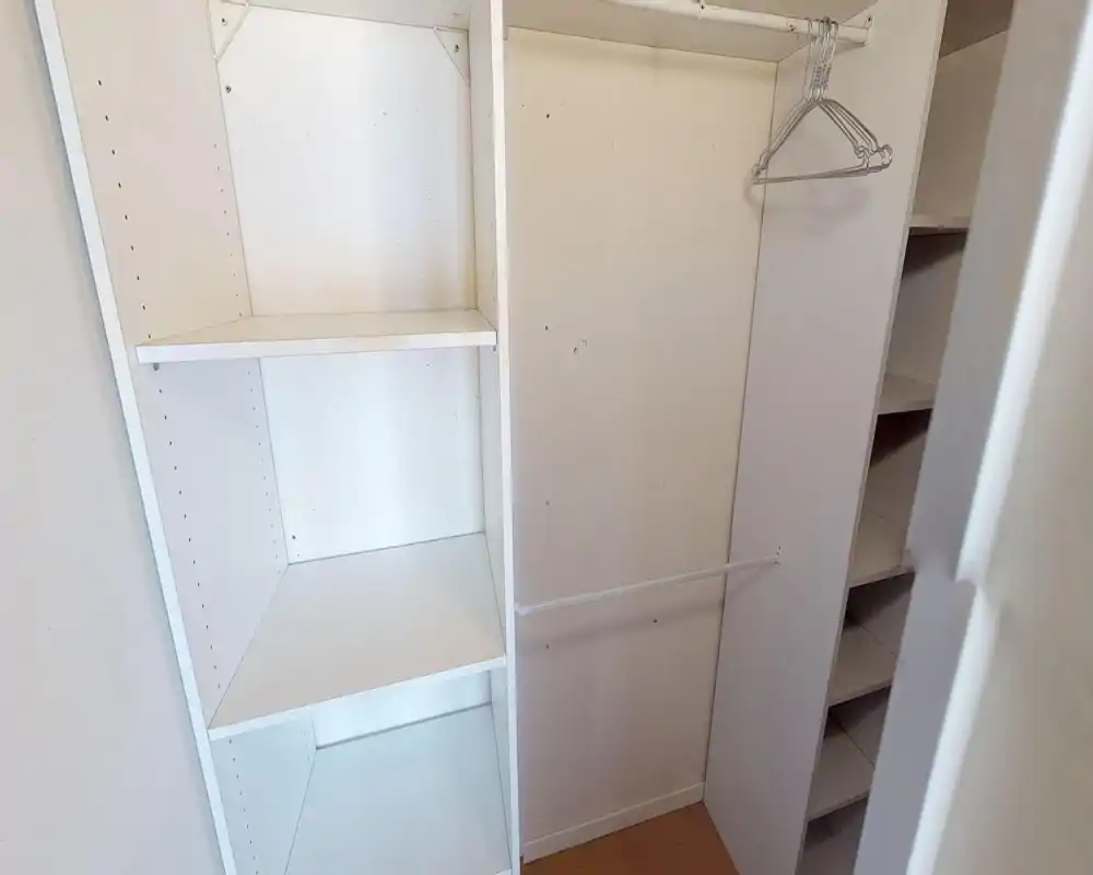 Petit dressing/entrée de rangement avec étagères blanches intégrées, tringle et cintres métalliques ; murs neutres et sol moquetté.