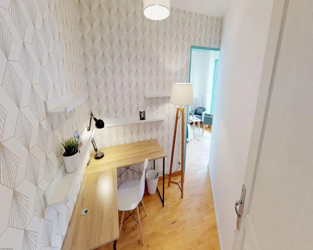 Coin bureau lumineux et compact intégré au salon : bureau en bois avec lampe, chaise blanche, papier peint décoratif et lampadaire près de l'ouverture donnant sur l'espace détente.