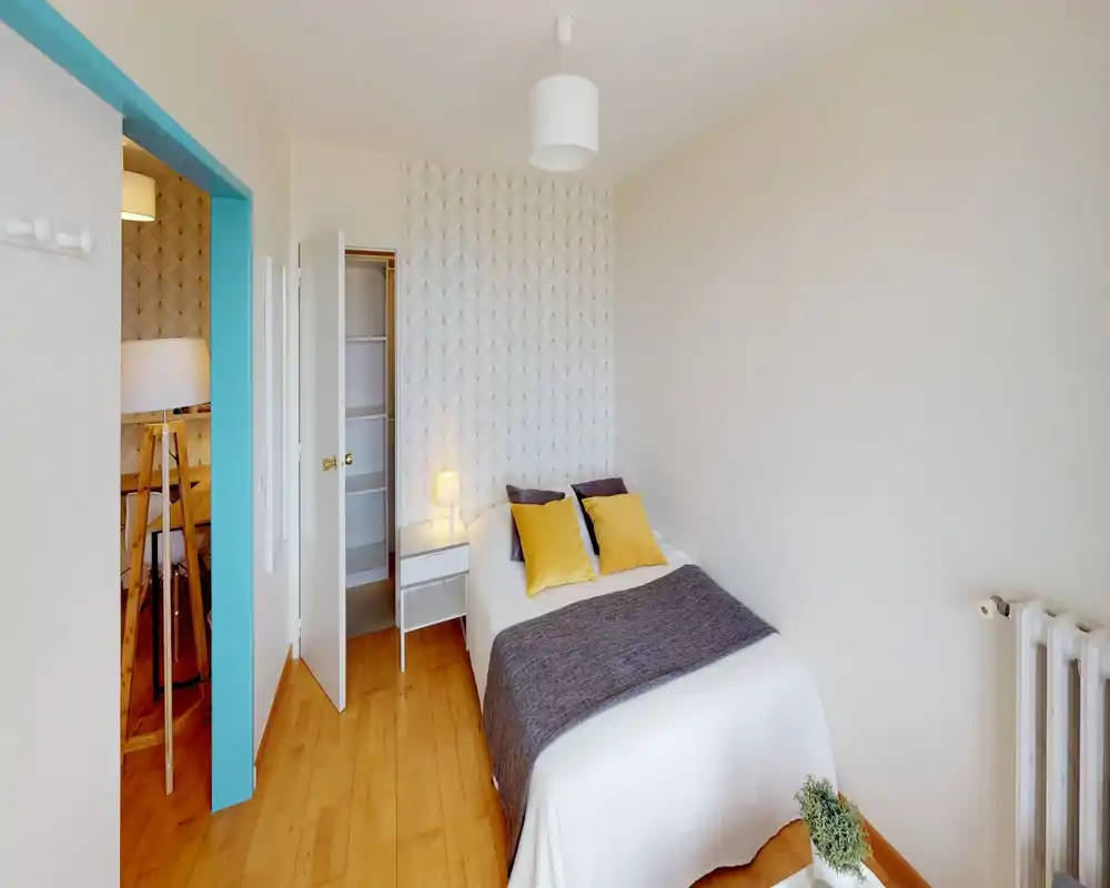 Chambre lumineuse et mise en scène avec un lit double central en linge blanc, coussins jaunes d'accent et mur texturé; table de chevet avec lampe et parquet apportent une ambiance chaleureuse et moderne.