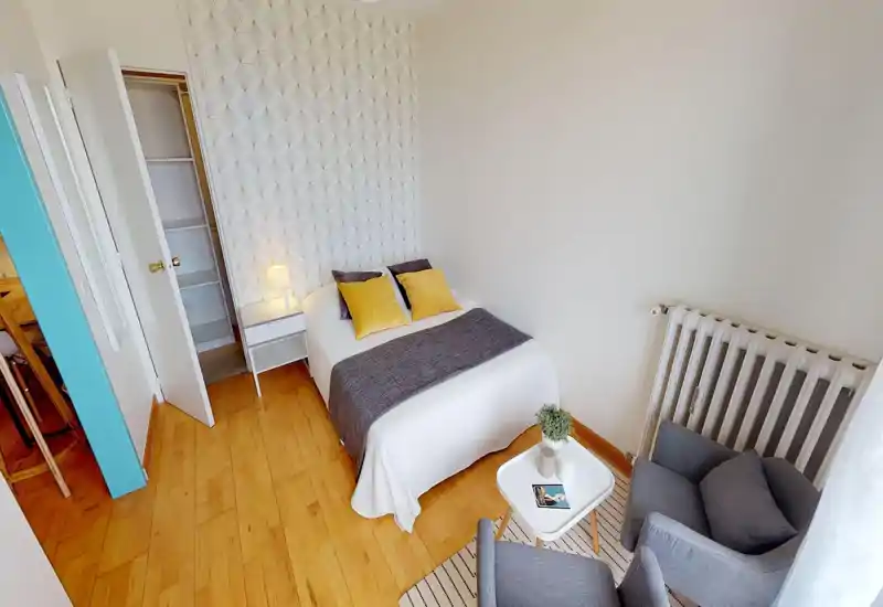 Chambre lumineuse et bien décorée avec un lit double en linge blanc, jeté gris et coussins jaunes ; table de chevet avec lampe et petit coin salon pour une ambiance chaleureuse.