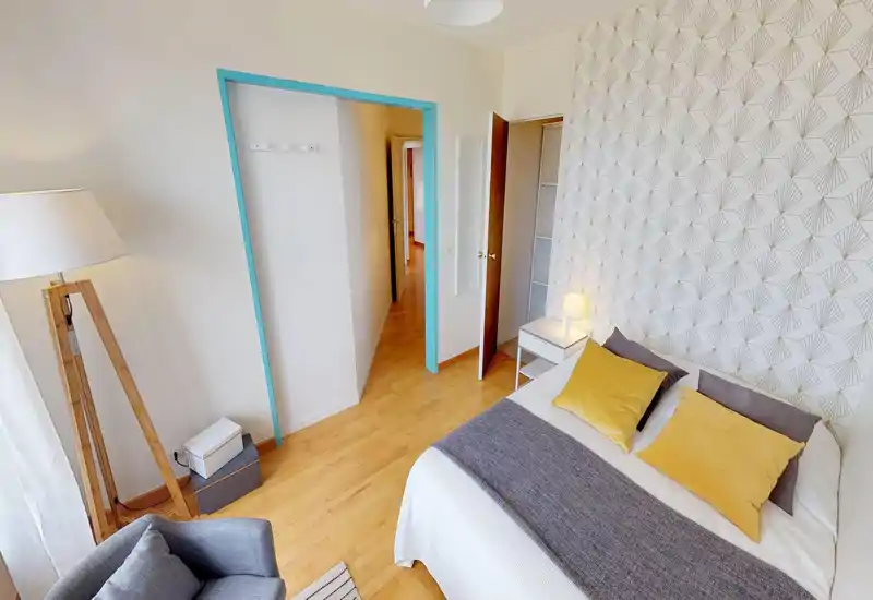 Chambre lumineuse et bien décorée avec un grand lit en linge neutre et coussins moutarde, table de chevet avec lampe, mur décoratif et un fauteuil près de la fenêtre.