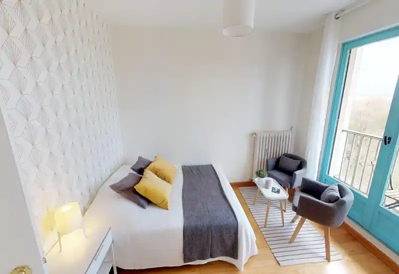 Chambre lumineuse et décorée avec un lit double en linge blanc, un jeté gris et des coussins jaunes, ainsi qu'un coin salon près d'une grande fenêtre donnant sur le balcon.