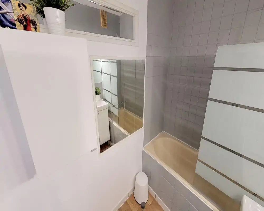 Salle de bains lumineuse et bien entretenue avec murs carrelés, baignoire encastrée et pare-douche en verre dépoli. Couleurs neutres et sol effet bois pour un espace propre et fonctionnel.
