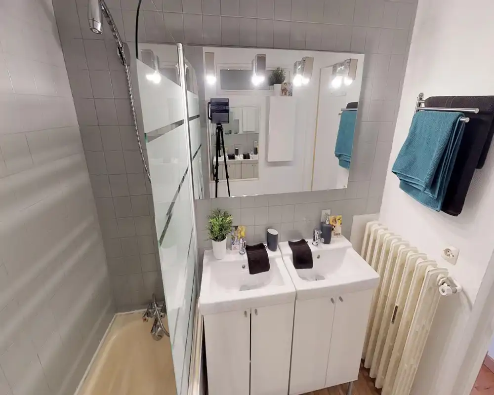 Salle de bain lumineuse et moderne avec double vasque, grand miroir, baignoire-douche avec paroi en verre et serviettes soigneusement pliées.