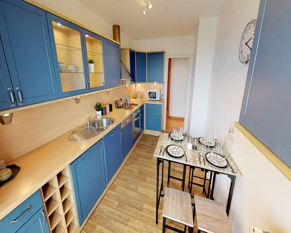 Cuisine moderne et lumineuse avec placards bleus, four encastré et évier, plus un petit coin repas dressé pour deux — un espace compact et bien agencé, parfait pour de courts séjours.