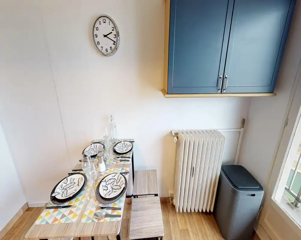 Coin repas compact avec table dressée pour quatre, sets de table géométriques et tabourets sous un placard mural ; belle lumière naturelle, radiateur et poubelle visibles.