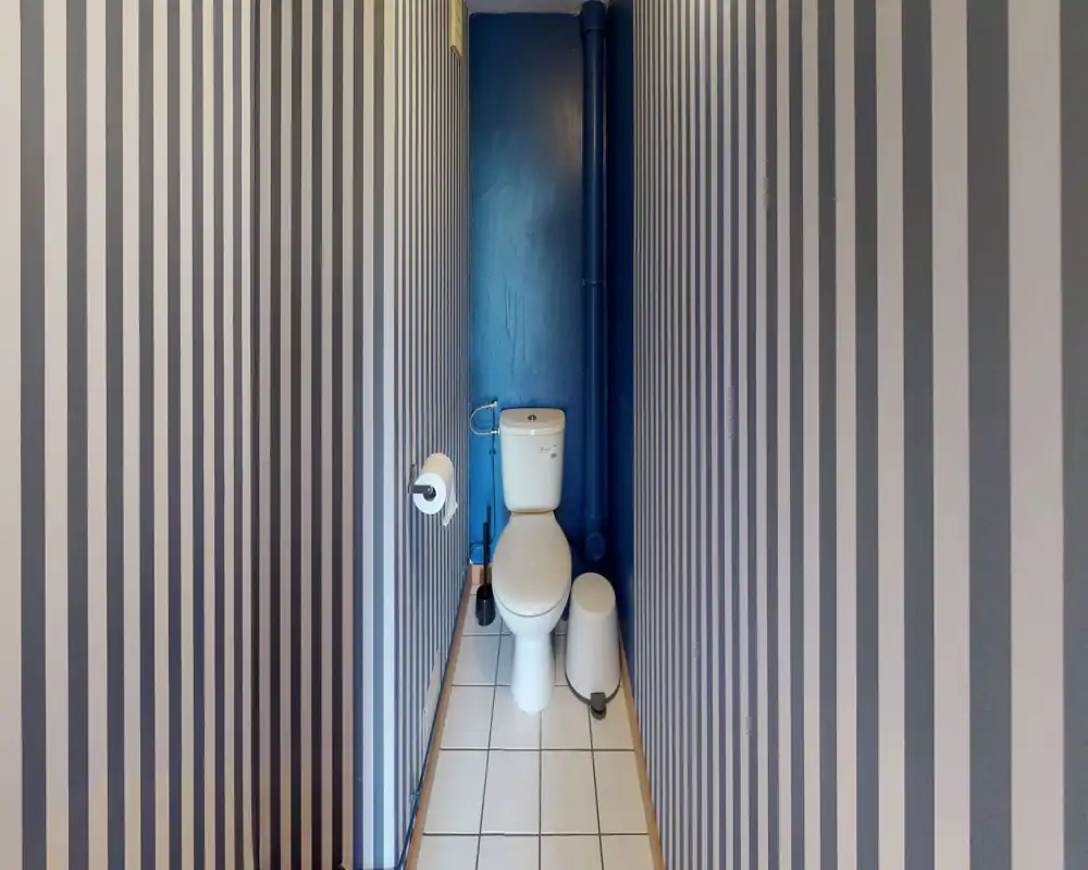 Petite pièce WC avec un WC blanc, sol carrelé et murs rayés bleus ; design simple et soigné, idéal comme toilettes invité.
