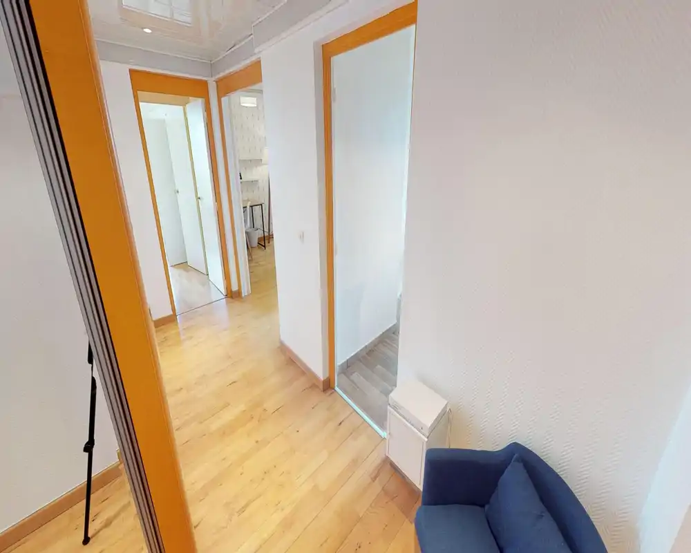 Espace de vie en longueur avec parquet, portes vers les pièces adjacentes et un fauteuil bleu ; lumineux, propre et minimaliste.