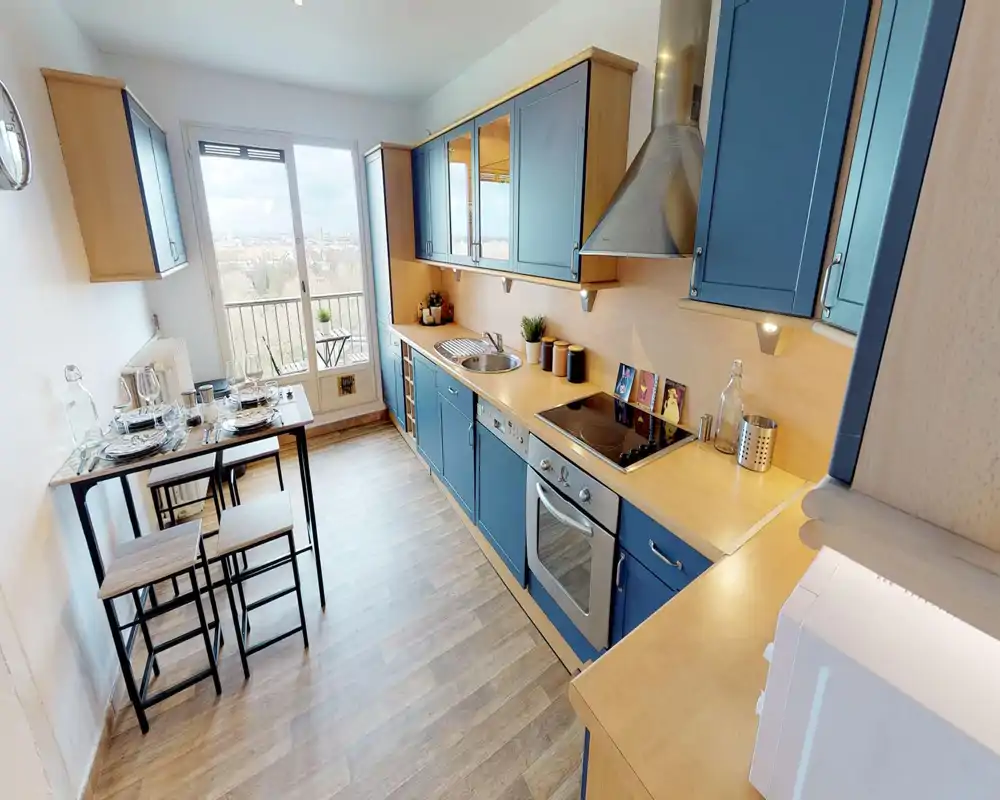 Cuisine moderne en enfilade avec meubles bleus, four encastré et plaque électrique, évier intégré sur plan de travail en bois clair et petit coin repas près de la fenêtre avec vue sur le balcon.