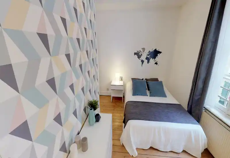 Camera da letto luminosa e ben arredata con letto matrimoniale in lenzuola bianche, cuscini blu e plaid scuro. Parete geometrica, comodino con lampada e pavimento in legno naturale danno un tocco fresco e accogliente.
