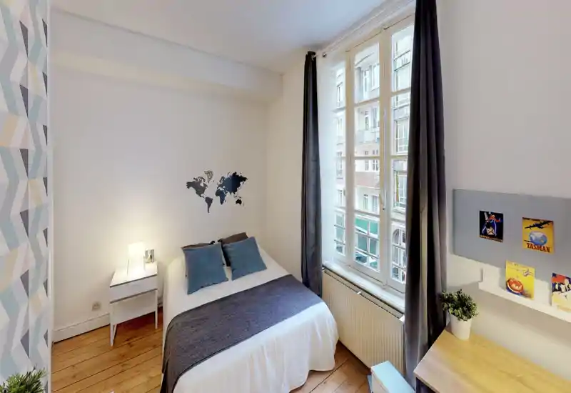 Camera da letto piccola e luminosa con letto matrimoniale in biancheria bianca e dettagli blu, comodino con lampada, ampia finestra con tende e una piccola scrivania — ambiente accogliente e moderno ideale per brevi soggiorni.