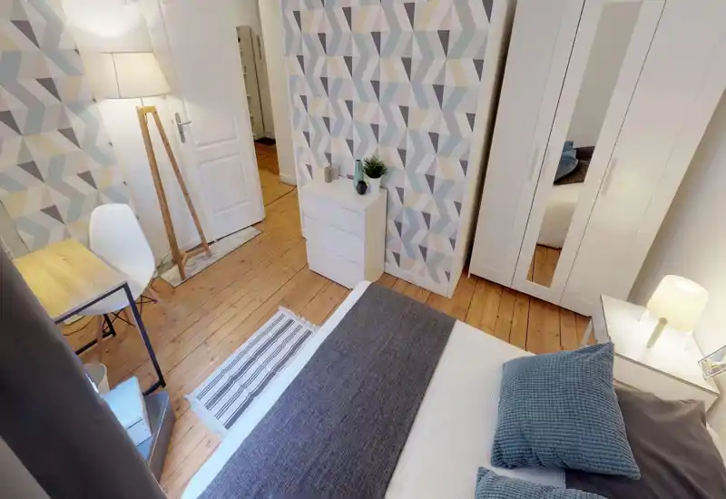 Camera da letto luminosa e curata con un letto matrimoniale in primo piano, cuscini decorativi, pavimento in legno e un armadio bianco con anta a specchio — ambiente accogliente e ben illuminato, ideale per annunci.