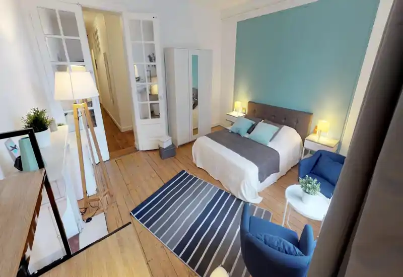 Ampia camera matrimoniale ben arredata con letto comodo, comodini e area relax vicino alla finestra. Pavimenti in legno chiaro e una parete d'accento color teal creano un'atmosfera moderna e rilassante, perfetta per affitti brevi.