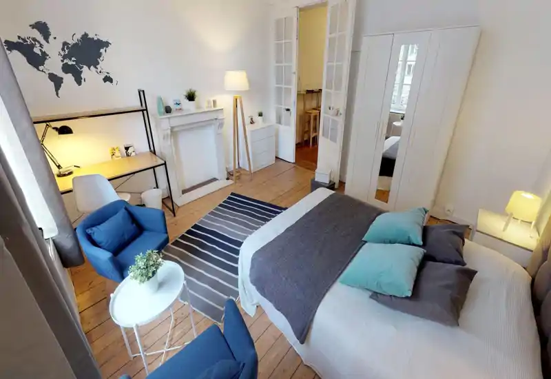 Camera da letto luminosa e di stile con letto matrimoniale ben rifinito, cuscini decorativi e lampada da comodino. La stanza presenta un armadio con specchio, un angolo salotto con poltrone blu e un tavolino, oltre a una scrivania e una mappa decorativa sulla parete.