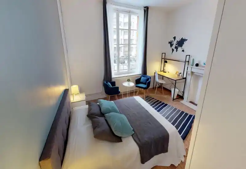 Camera da letto luminosa e ben arredata con un grande letto in primo piano, due poltroncine vicino alla finestra e una scrivania compatta — ideale per riposare e lavorare.