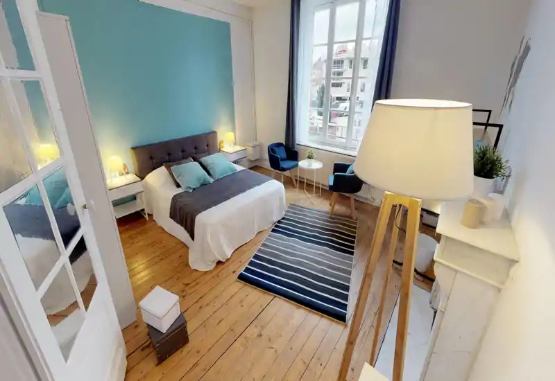 Camera da letto luminosa e elegante con letto matrimoniale, comodini e un'area salotto vicino a una grande finestra. Pavimenti in legno, cuscini coordinati e luci calde creano un'atmosfera accogliente.