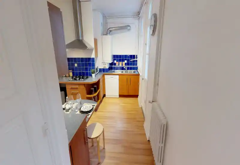 Cuisine en enfilade compacte et lumineuse avec meubles en bois, crédence en carreaux bleus, plaque à gaz et évier — idéale pour un petit appartement.
