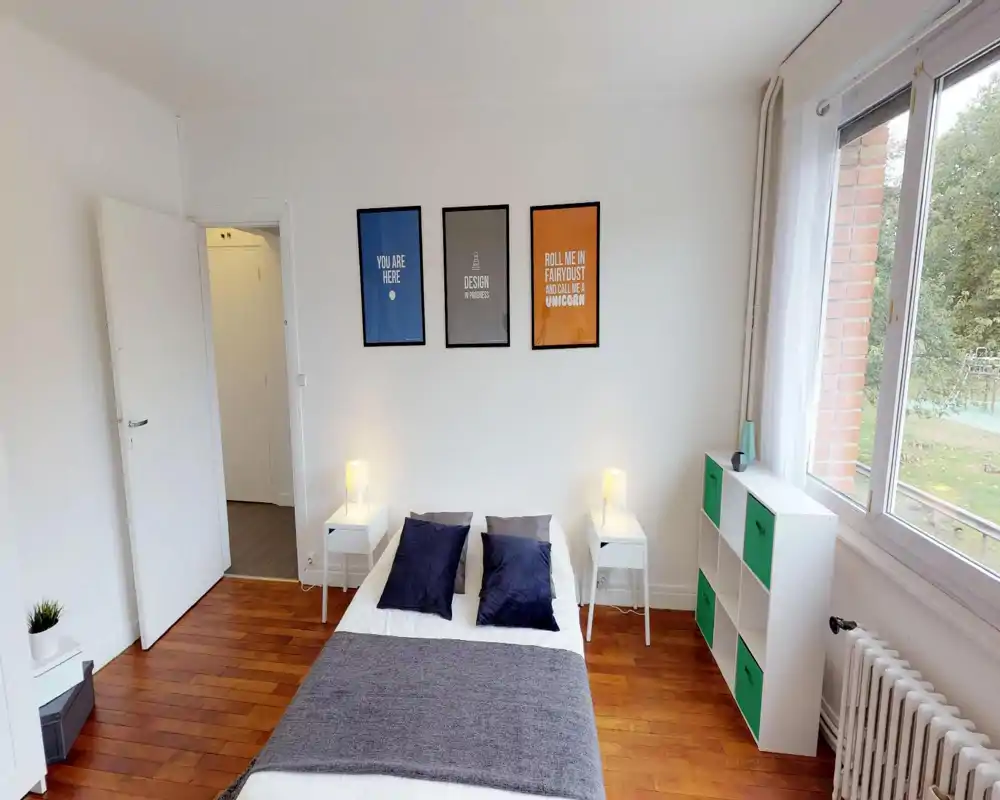 Chambre lumineuse et soignée avec un lit double centré contre un mur blanc, deux tables de chevet avec lampes, coussins décoratifs et une grande fenêtre offrant lumière naturelle et vue sur la verdure.