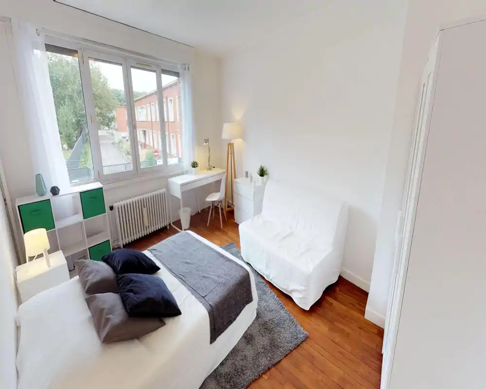 Chambre lumineuse et bien mise en scène avec lit double, textiles confortables et une grande fenêtre apportant beaucoup de lumière naturelle. La pièce comprend un bureau compact, des rangements et un petit canapé blanc, idéale pour un séjour minimaliste et agréable.