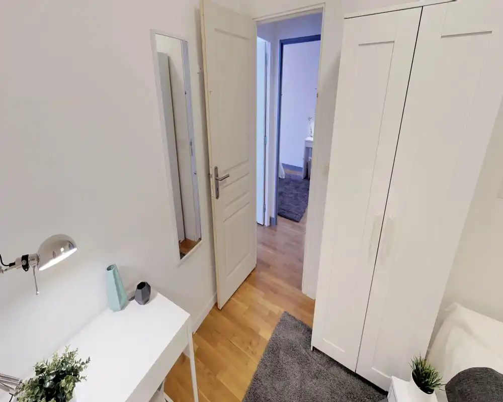 Angolo di camera arredato in stile minimal: armadio bianco, parte del letto visibile sul bordo destro, scrivania con lampada e piante decorative — luminoso e ordinato.