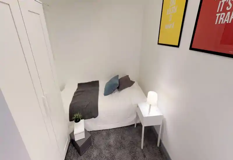 Piccola camera singola ben arredata con letto ordinato, comodino con lampada, armadio e quadri a parete. Luminosa, minimale e accogliente — ideale per un viaggiatore singolo o un ospite.