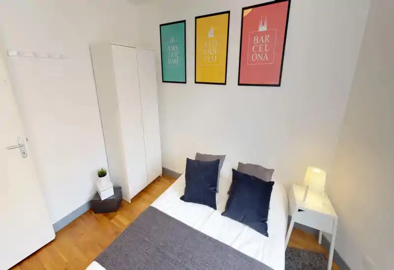 Bright, neatly staged bedroom with a double bed dressed in white linens, decorative navy cushions and a grey throw; a wardrobe and bedside table with lamp complete the simple, modern layout.