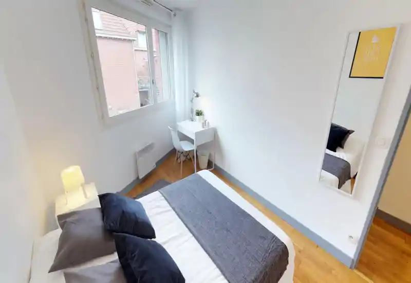 Bright, compact bedroom with a double bed dressed in neutral linens, dark accent pillows and a small white desk by the window — clean minimalist styling and warm wood floors.