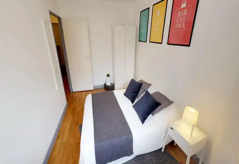 Bright, neatly styled bedroom with a double bed dressed in white linens, navy cushions and a gray runner; bedside table with lamp and a slim wardrobe optimize the compact space.