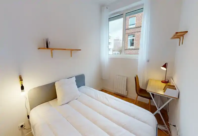 Chambre lumineuse et minimaliste avec un lit double confortable, une lampe de chevet et un petit bureau près de la fenêtre — idéale pour un séjour cosy et pratique.