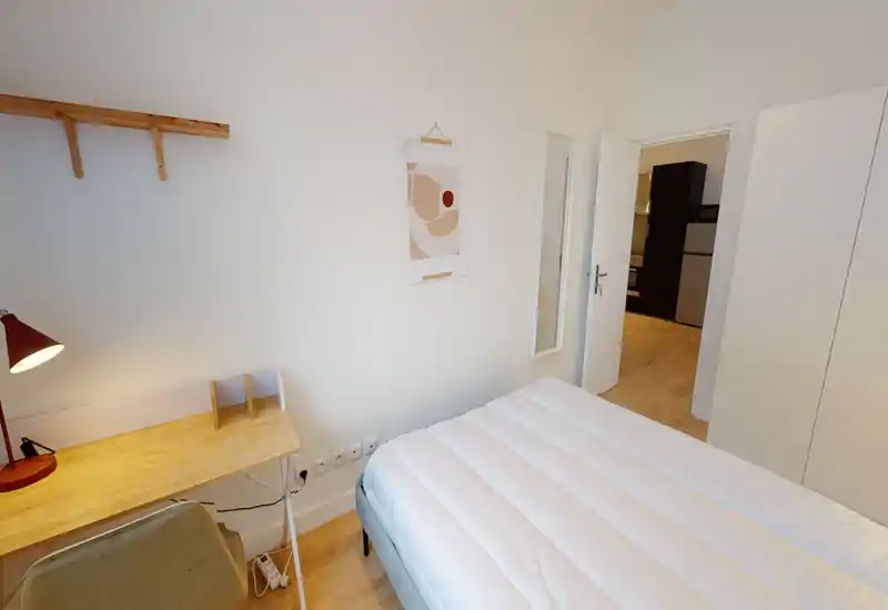 Chambre lumineuse et minimaliste avec un lit double bien fait, un bureau en bois avec lampe et une armoire près de la porte ; style épuré et moderne, idéal pour une location courte durée.