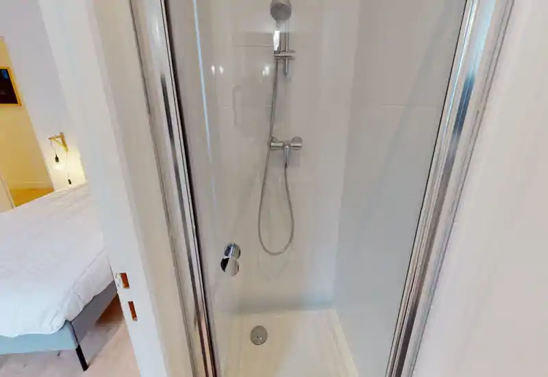 Compact tiled shower with glass door and chrome fixtures; clean, bright cabin-style bathroom visible from adjacent bedroom.
