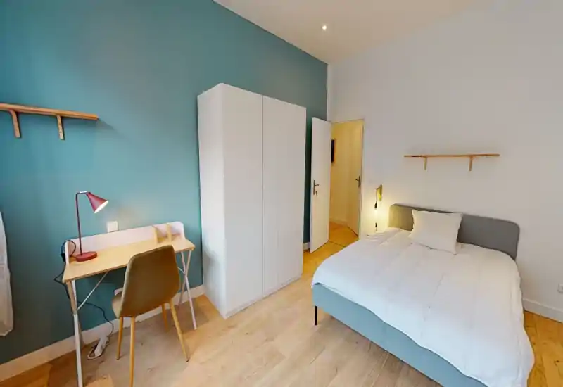 Bright, modern bedroom with a comfortable double bed, soft white bedding and a minimalist wardrobe. A small desk with lamp and a cozy chair sits against a teal accent wall — ideal for work and relaxation.