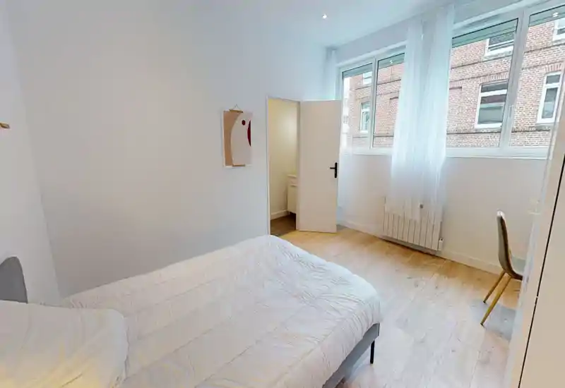Bright, minimal bedroom with a comfortable double bed, large windows bringing natural light, and simple desk seating—ideal for a restful stay.